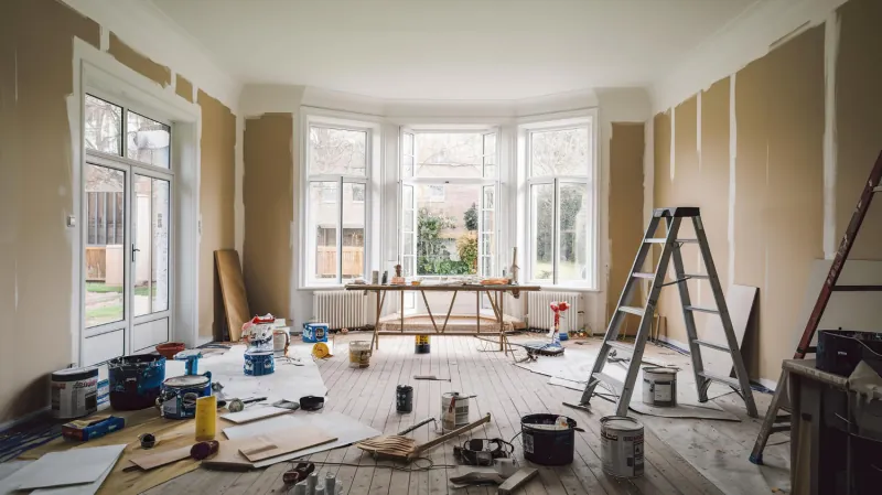 Living room mid renovation with a stepladder, drop cloths and open paint cans across a bare timber floor