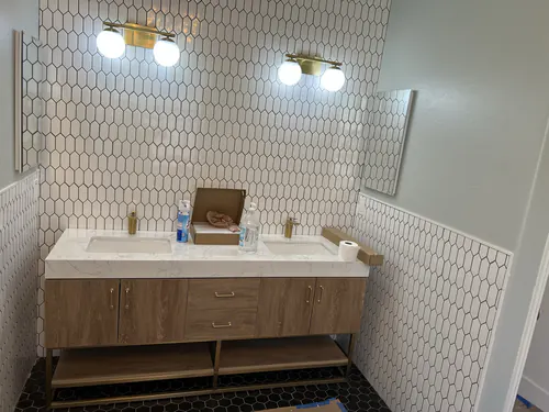 Double vanity with a light wood cabinet, stone top, two brass globe sconces and white hexagon wall tile behind