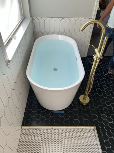 Freestanding soaking tub set on black hexagon floor tile with a brass floor mounted tub filler beside it