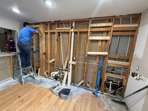 The same wall stripped back to bare studs with the drain stack and wiring exposed and a carpenter working from a step ladder