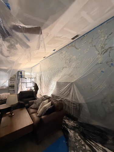 Living room ceiling scraped back to bare drywall with the tape seams showing, and the walls and floor still covered in plastic