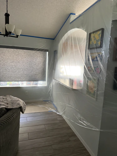 Living room wall and the arched kitchen opening covered in plastic sheeting, under a popcorn ceiling taped at the edges
